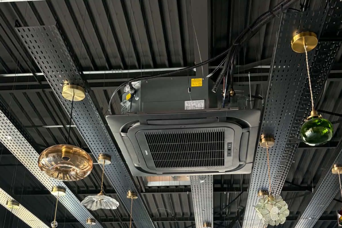 Cassette de climatisation suspendue sous une charpente métallique, entre les chemins de câbles et les suspensions